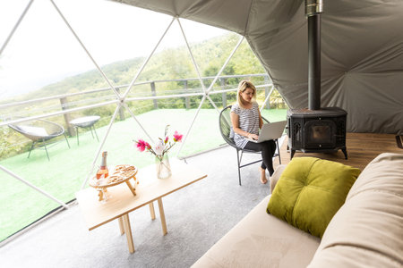 middle aged woman uses a laptop resting and spending time at Glamping house on holidays. holiday dome tent. Cozy, camping, hygge, lifestyle conceptの写真素材