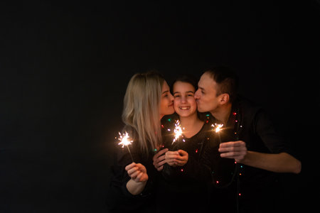 Happy family in black - woman, man and little girl, with a sparkler in hand the New Years Christmas at home. The girl at the father on hands. New year 2023.の写真素材