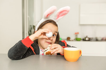 Curious small girl in bunny ears peeking out from behind kitchen table with tasty easter cake and looking at camera with surprise. Focus on childの写真素材
