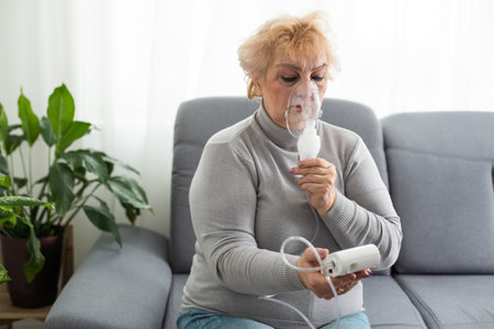 Sick senior woman has a viral infection or flu, labored breathing, asthma. An elderly female is using inhaler, nebulizer for self treatment sitting alone on the sofa at homeの写真素材