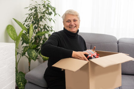 Smiling elderly woman customer receive post shipment parcel at home, happy old senior grandma hold open cardboard box sit on sofa in living room, online shopping order fast courier delivery concept.の写真素材