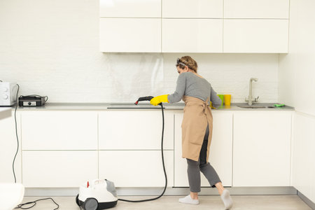 Cropped photo of a woman in uniform cleaning filters for kitchen range hood with steam cleaner while her male colleague working on the background. Cleaning services concept. Housework.の写真素材