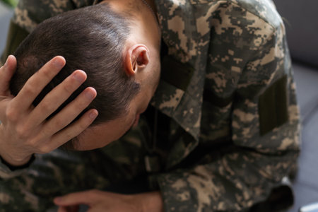 Unhappy man in camouflage uniform sitting at arm chair over white studio background, touching his head, soldier suffering from posttraumatic stress disorder, returning back home after military serviceの写真素材