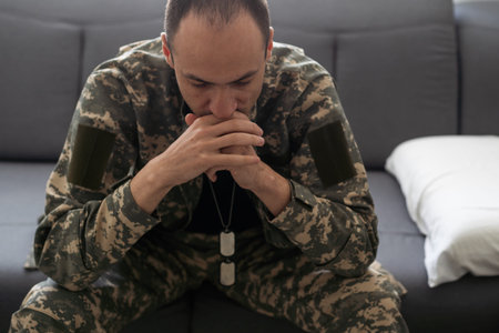 Serious anxious and nervous military man suffers from depression while sitting alone at home. Pensive man in military uniform sitting on sofa propping his head with his hands. PTSD concept.の写真素材