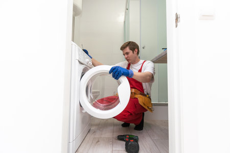 The young handsome repairman in worker suit with the professional tools box is fixing the washing machine in the bathroom.の写真素材