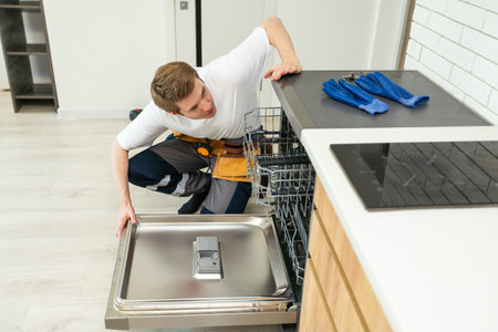 Technician or worker in uniform installs dishwasher into the kitchen furniture. Repairman wear worker suit repairing maintenance of dishwasher. Master in protective gloves fix dishwasher.の写真素材