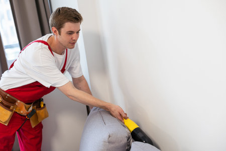 Rear View Of Young Male Worker Cleaning Sofa With Vacuum Cleaner