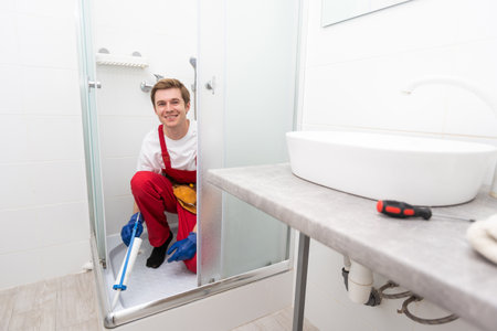 Young man in uniform repairs the shower door in the bathroom. A male repairman repairs the shower cabin.の写真素材