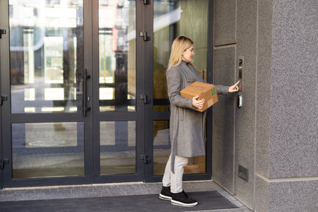 Happy woman ringing on doorbell at building entrance. Using intercom