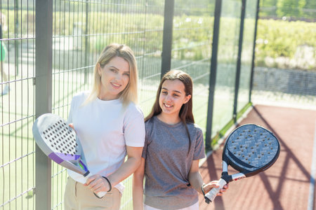 positive mother and daughter standing on court with padel rackets during break and looking at camera.の写真素材