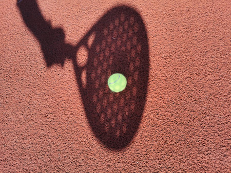 High angle of yellow ball placed on court and covered with shadow of crop person holding padel racketの写真素材