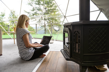 Woman working on laptop geo dome tents. Green, blue background. Cozy, camping, glamping, holiday, vacation lifestyle concept. Outdoors cabin, scenic background.の写真素材