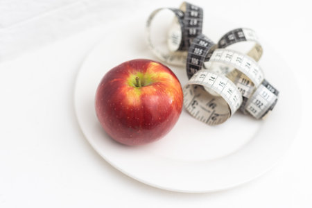 Slimming. A red-orange apple wrapped in a measuring tape lies on a white plate, on a white background. Calls to lose weightの写真素材