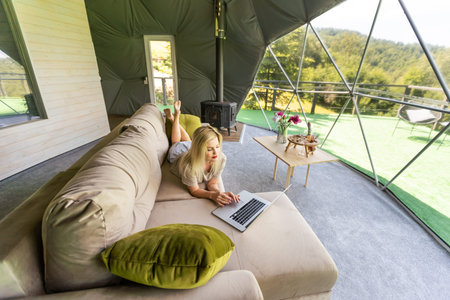 Young caucasian woman using and watching on laptop computer on in dome camping hotel. Tourism, vacation and weekend.の写真素材