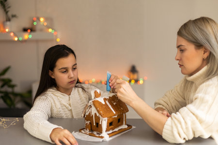 mother and daughter decorating gingerbread house. Beautiful living room with lights. Happy family celebrating holiday together.の写真素材