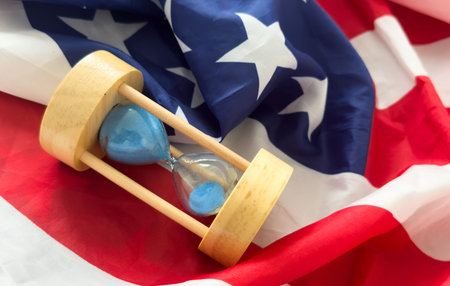 Hourglass on table with USA flag in the background. Sand glass on old wooden table with American...
