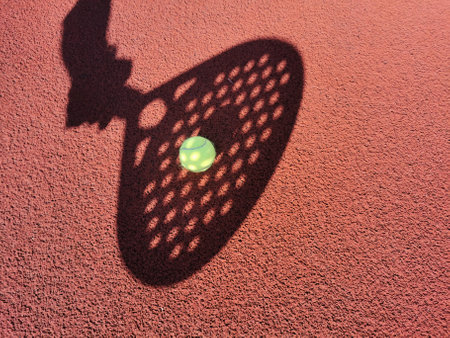 High angle of yellow ball placed on court and covered with shadow of crop person holding padel racketの写真素材