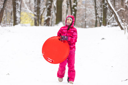 Pretty little girl wearing pink jacket and knitted hat walking in snowy winter park. Child playing with ice skating plate in forest. Family vacation with child in mountainsの写真素材