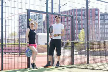Portrait of positive young woman and adult man standing on padel tennis court, holding racket and ball, smilingの写真素材