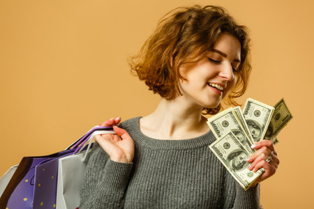 Photo of cheerful young woman holding fan of money and colorful shopping bags, isolated on yellow backgroundの写真素材
