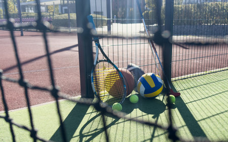 A collection of sports equipment including tennis balls, a tennis racket, and a volleyball on a court surface viewed through nettingの写真素材