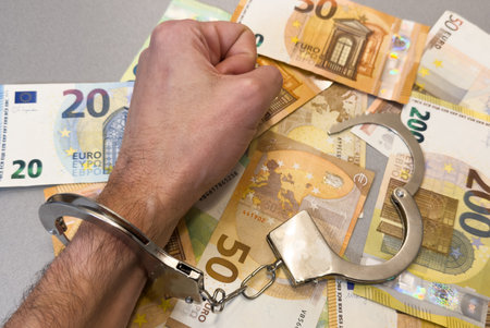 Folded hands and handcuffed hands resting on a table in front of money.の写真素材