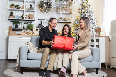 festive girl openng xmas gifts on Christmas eve with mom and dad, decorated kitchen interior, copy spaceの写真素材
