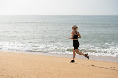 Woman jogging on a beach, enjoying the ocean view. Beach running, ocean breeze, and fitness are key. Active lifestyle by the sea, embracing the beach vibes. Woman exercising at a beach.の写真素材