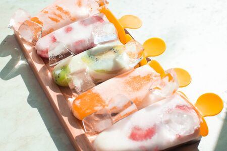 set assortment of homemade ice cream on a stick with pieces of fruit on ice on a wooden tray on a light backgroundの写真素材