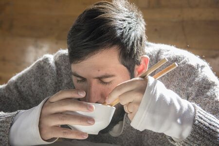 Portrait of a bearded man seating and eating from a bowl ,using chopsticks, in a wooden cabin.の写真素材