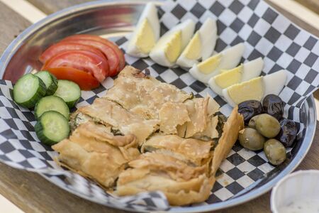 Top view of a plate, with homemade traditional turkish style Tabla Borek, boiled eggs, yogurt, sliced vegetables and olives, over a black and white paper napkin.の写真素材
