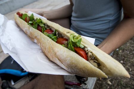 Woman holding large baguette sandwich with tomatoes, Maquis cheese, caper shrubs and basil, outdoors in the street, Corsicaの写真素材