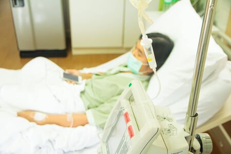 Patient lies in hospital bed with drip equipment in foreground. Focus is on the dripの写真素材