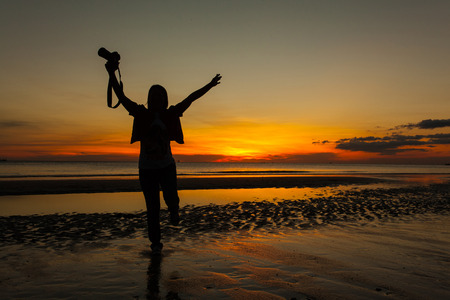 Silhouette woman photographer walking on beach with hand holding camera at sunrise.の写真素材