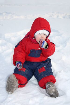 Little girl wearing warm jumpsuit sits on snow and eats snow at winterの写真素材