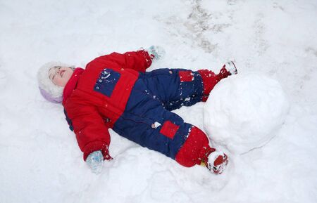 Little tired girl wearing warm jumpsuit lies on snow near big snowballの写真素材