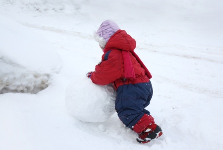 Back of little girl wearing warm jumpsuit is rolling big snowballの写真素材