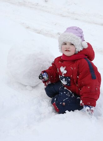 Little tired girl wearing warm jumpsuit sits on snow near big snowballの写真素材