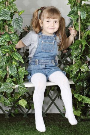 Happy little girl in denim jumpsuit sits on swing under green ivy の写真素材
