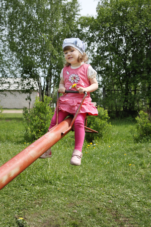 Happy little cute girl in pink rides on red seesaw at summer day.の写真素材