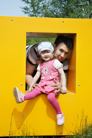 Happy little girl with father sits in window of yellow box at playground at summer day. の写真素材