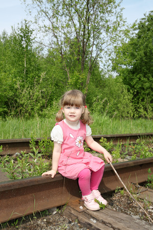 Little cute girl in pink dress with stick sits on old railway at summer day.  の写真素材