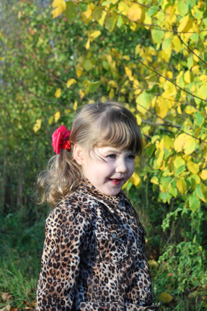 Smiling little girl looks away next to yellow trees in park at sunny autumn day.の写真素材
