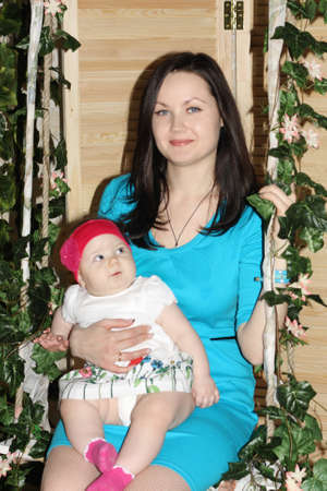 Beautiful woman in blue with baby sits on swing overgrown with green ivy.の写真素材