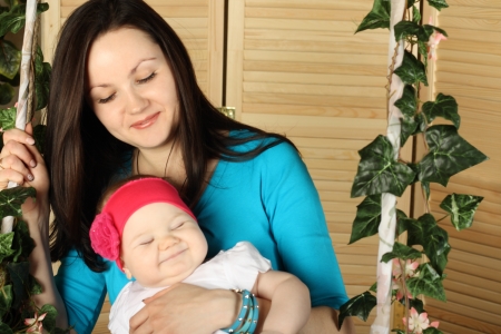 Beautiful woman in blue with smiling baby sits on swing overgrown with green ivy.の写真素材