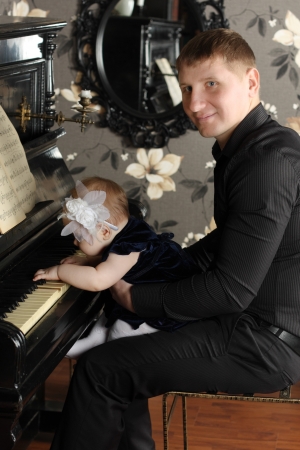 Smiling man in black with cute baby sits at piano in room. Baby plays piano.の写真素材