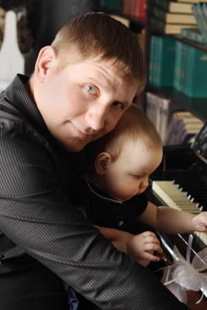 Cute baby sits at knees of her father and touches piano in room. Focus on baby.の写真素材