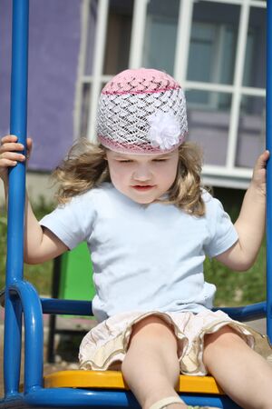 Little happy girl sits on swing at playground violet building at sunny day.の写真素材