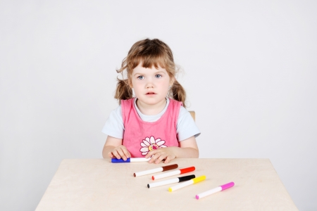 Little cute girl sits at wooden table with felt-tip pens on grey background.の写真素材
