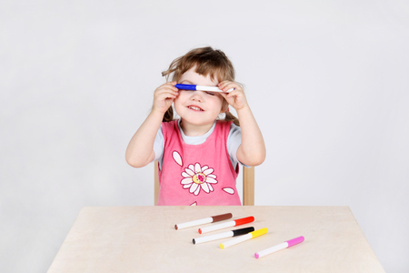 Little cute girl sits at wooden table and plays with felt-tip pens on grey background.の写真素材